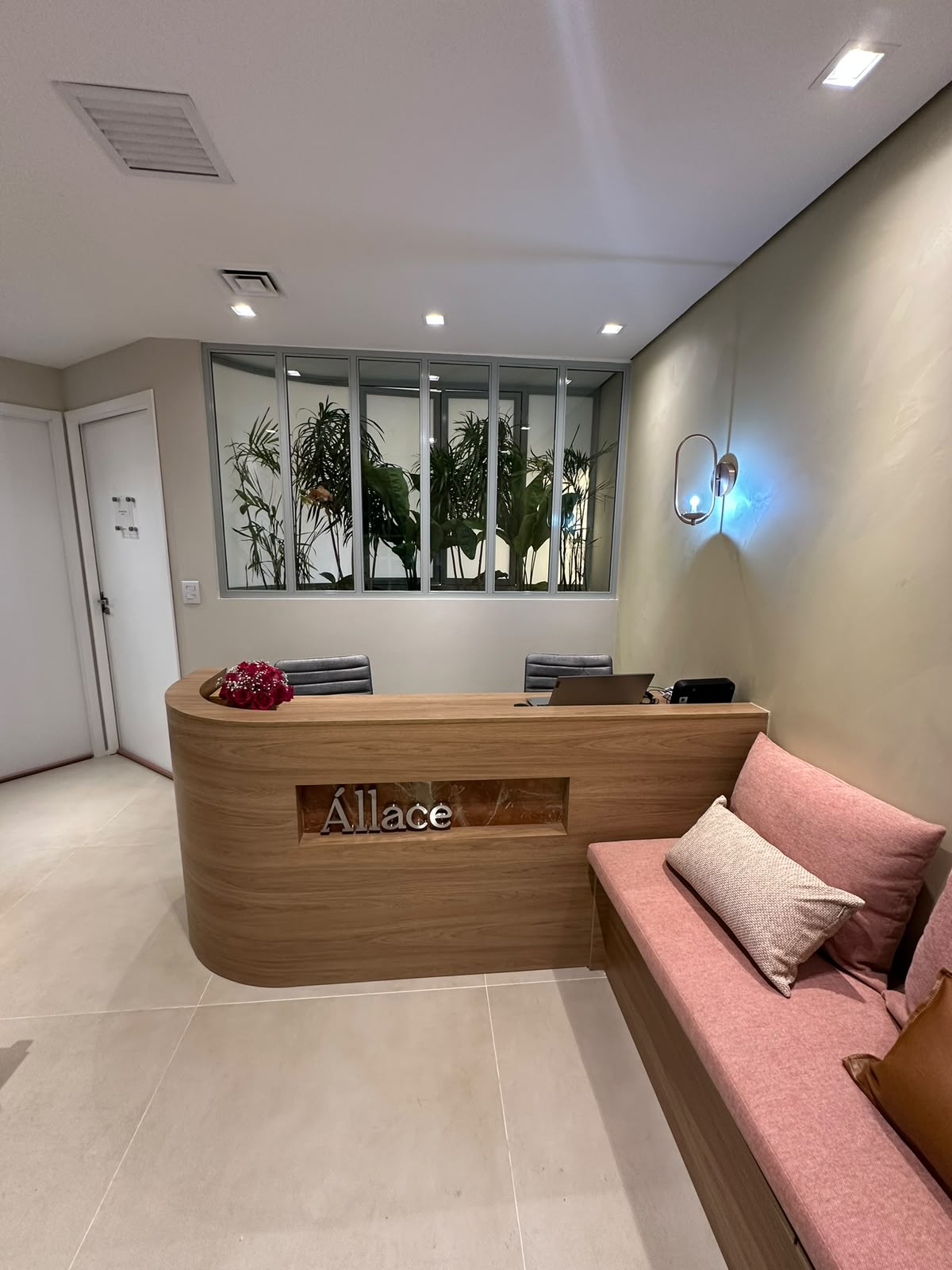 Recepção da Clínica Állace, com balcão de madeira, plantas ao fundo e sofá rosa